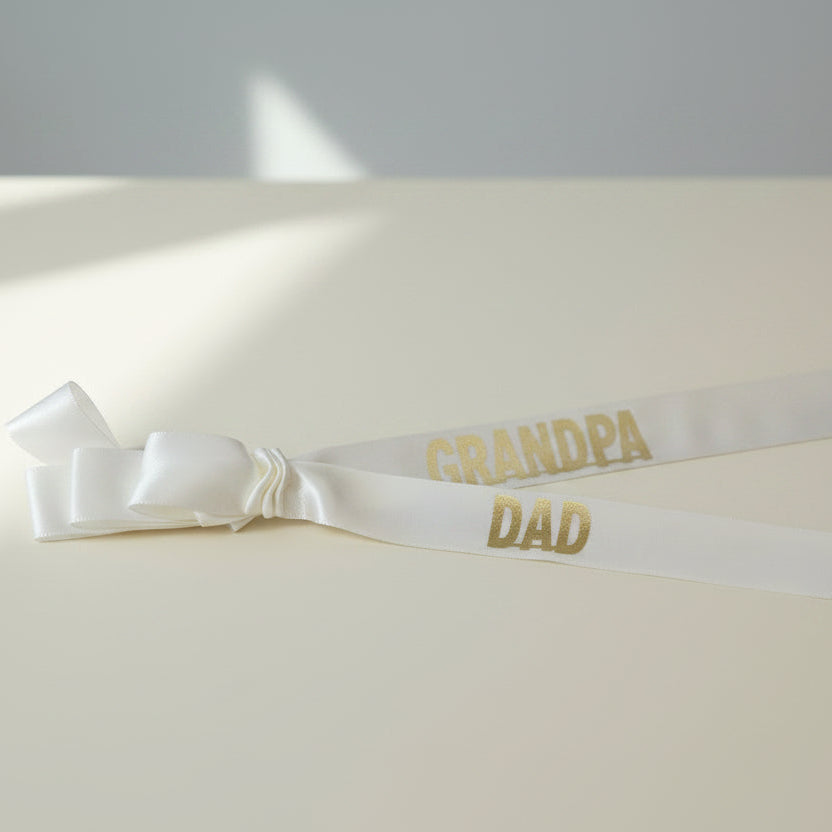 A white satin ribbon tied in a bow, printed with 'GRANDPA DAD' in gold lettering, laid flat on a wooden surface.